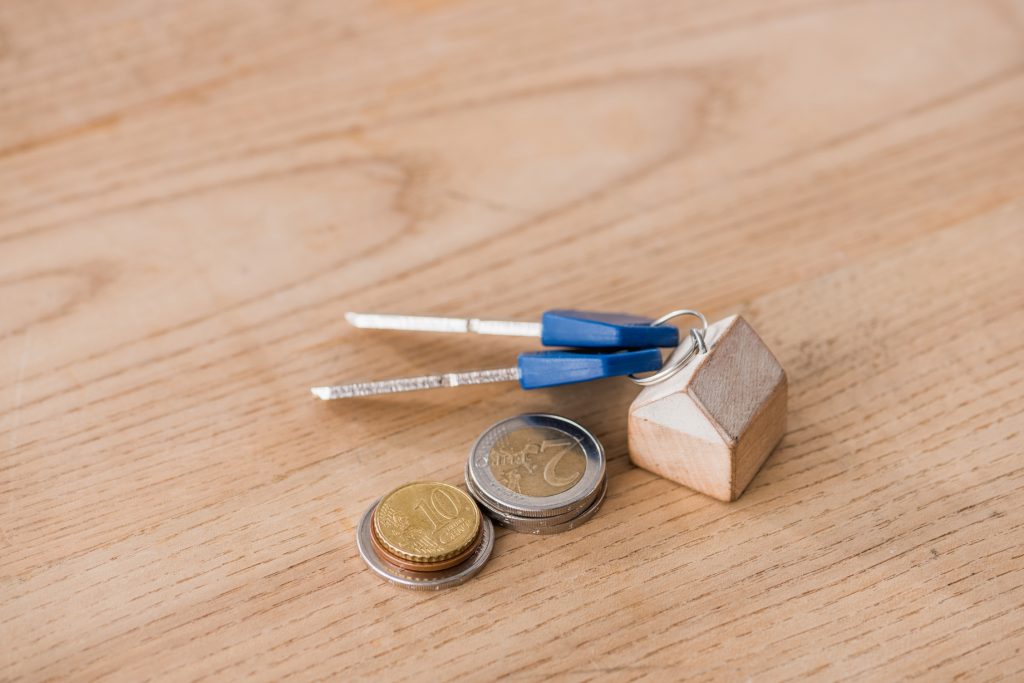 Keys With Toy House Trinket Near Golden And Silver Coins On Wooden Table